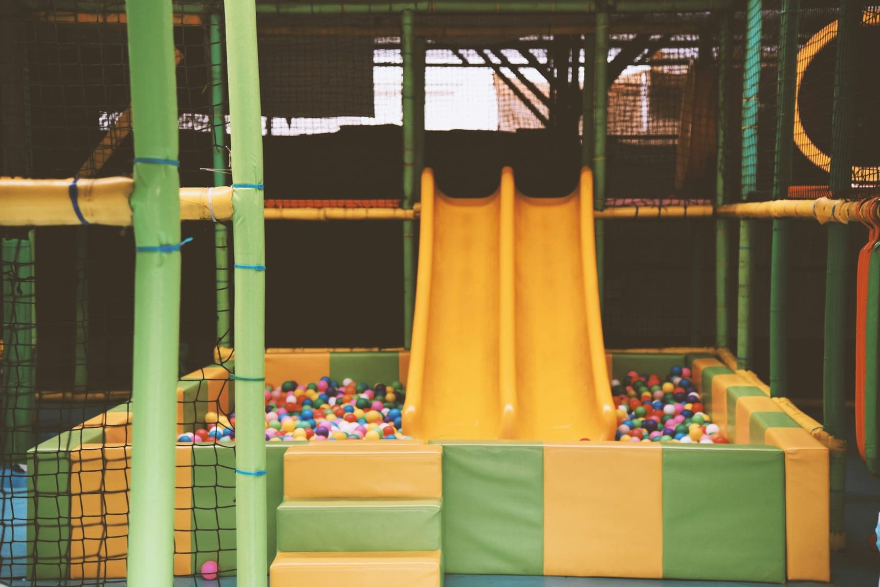 An indoor play structure: netting, padded posts, and a yellow twin slide into a ball pit